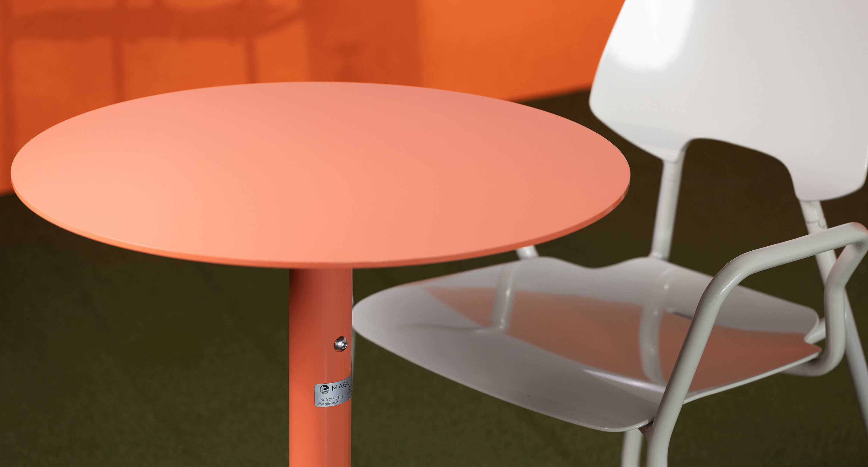 maglin_mtb-1900-ogden-cafe-tables-3 A round, coral-colored table is next to a white metal chair with a contoured seat on a green floor with an orange background.