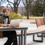 A person sits at an outdoor bench with a laptop, wearing headphones around their neck. Trees and a building labeled
