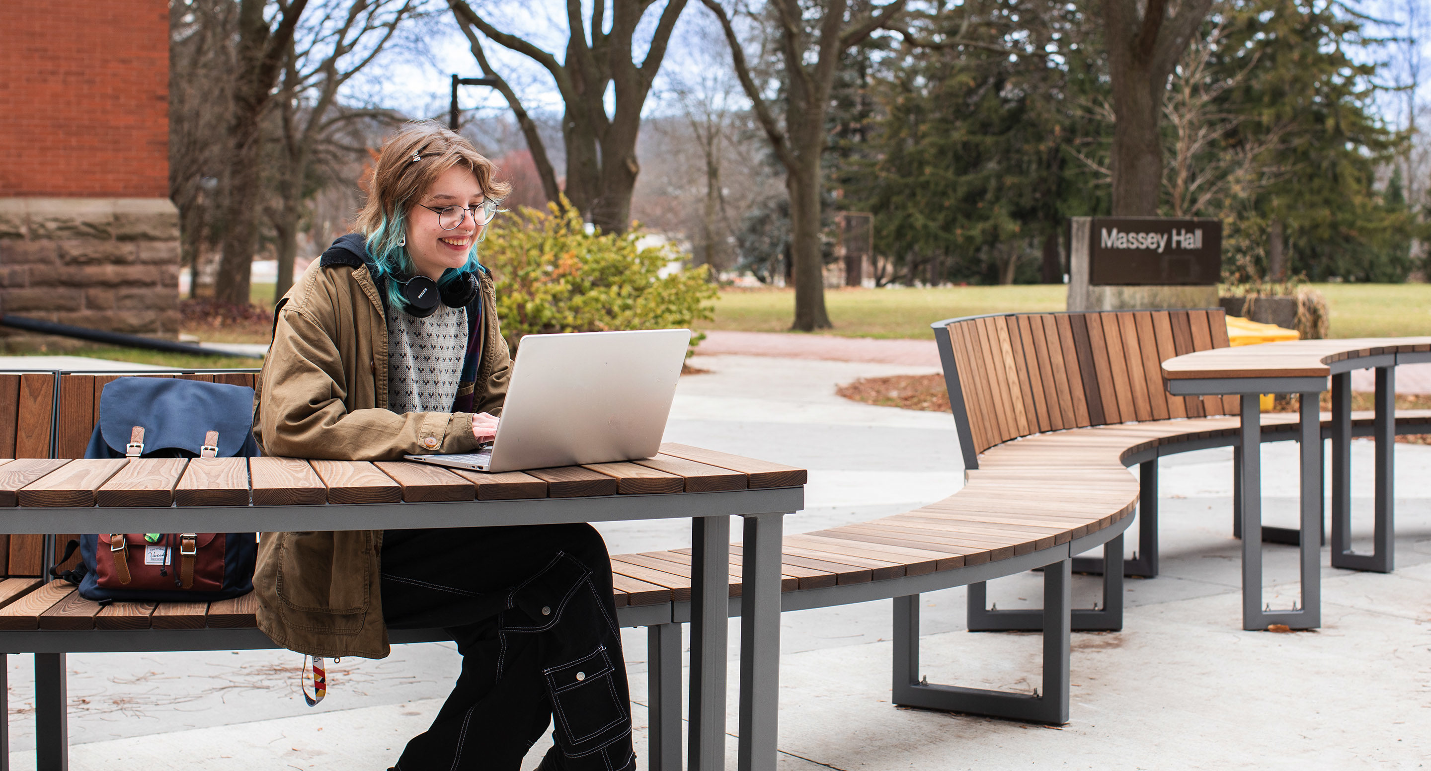 maglin_mtb-1900-ogden-modular-tables-1 A person sits at an outdoor bench with a laptop, wearing headphones around their neck. Trees and a building labeled