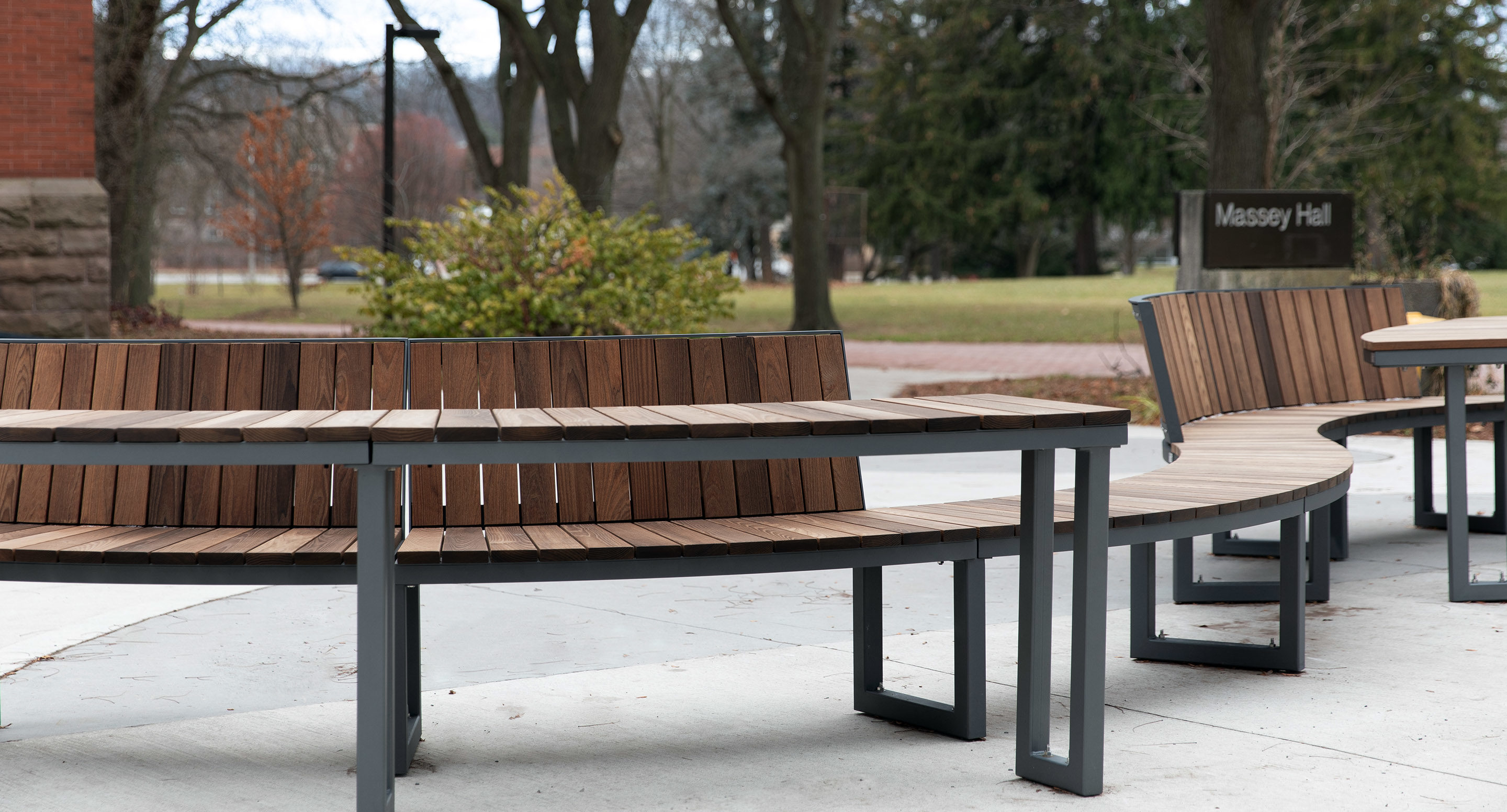 maglin_mtb-1900-ogden-modular-tables-2 Curved wooden benches with metal frames are arranged outdoors on a concrete surface near Massey Hall, surrounded by trees and grass.