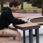 A person sits on a curved outdoor bench in a park, looking at a smartphone with both hands. Trees and grass are visible in the background.