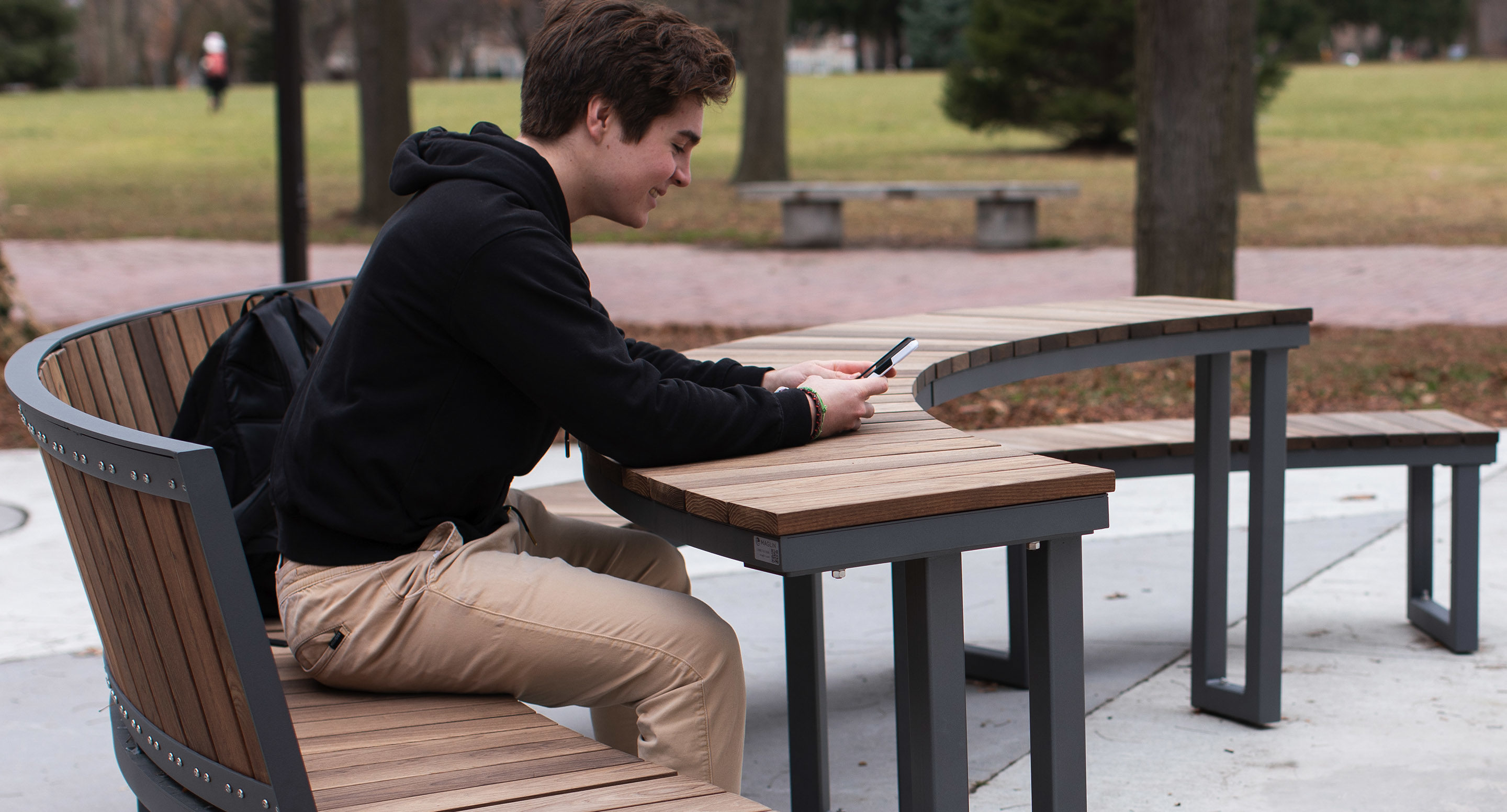 maglin_mtb-1900-ogden-modular-tables-3 A person sits on a curved outdoor bench in a park, looking at a smartphone with both hands. Trees and grass are visible in the background.