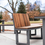 Outdoor seating area with wooden benches and tables on a concrete surface, surrounded by trees and fallen leaves in a park setting.