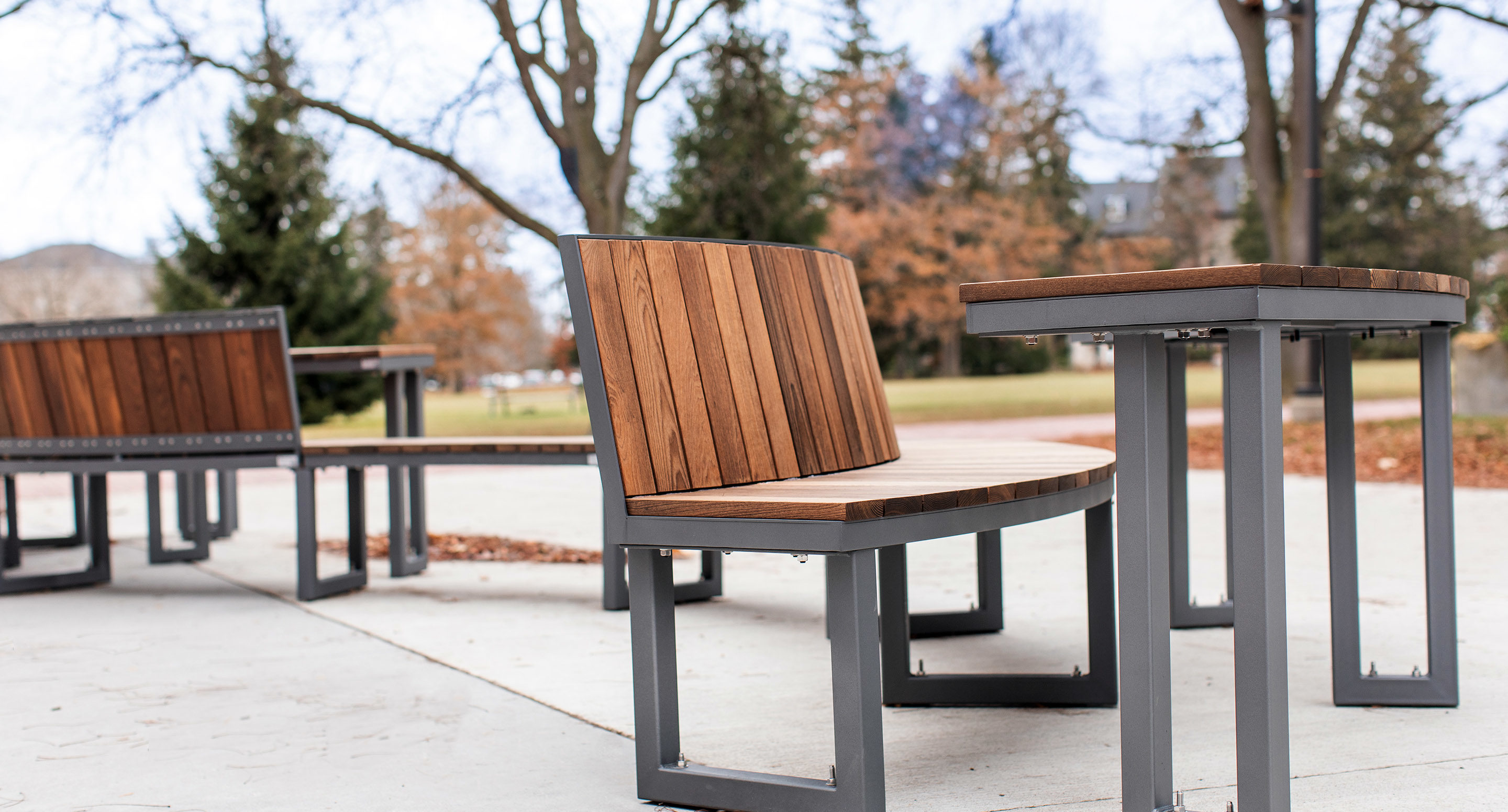 maglin_mtb-1900-ogden-modular-tables-4 Outdoor seating area with wooden benches and tables on a concrete surface, surrounded by trees and fallen leaves in a park setting.