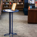A small round table with a framed document and a notebook stands in a library near bookshelves and a staircase.