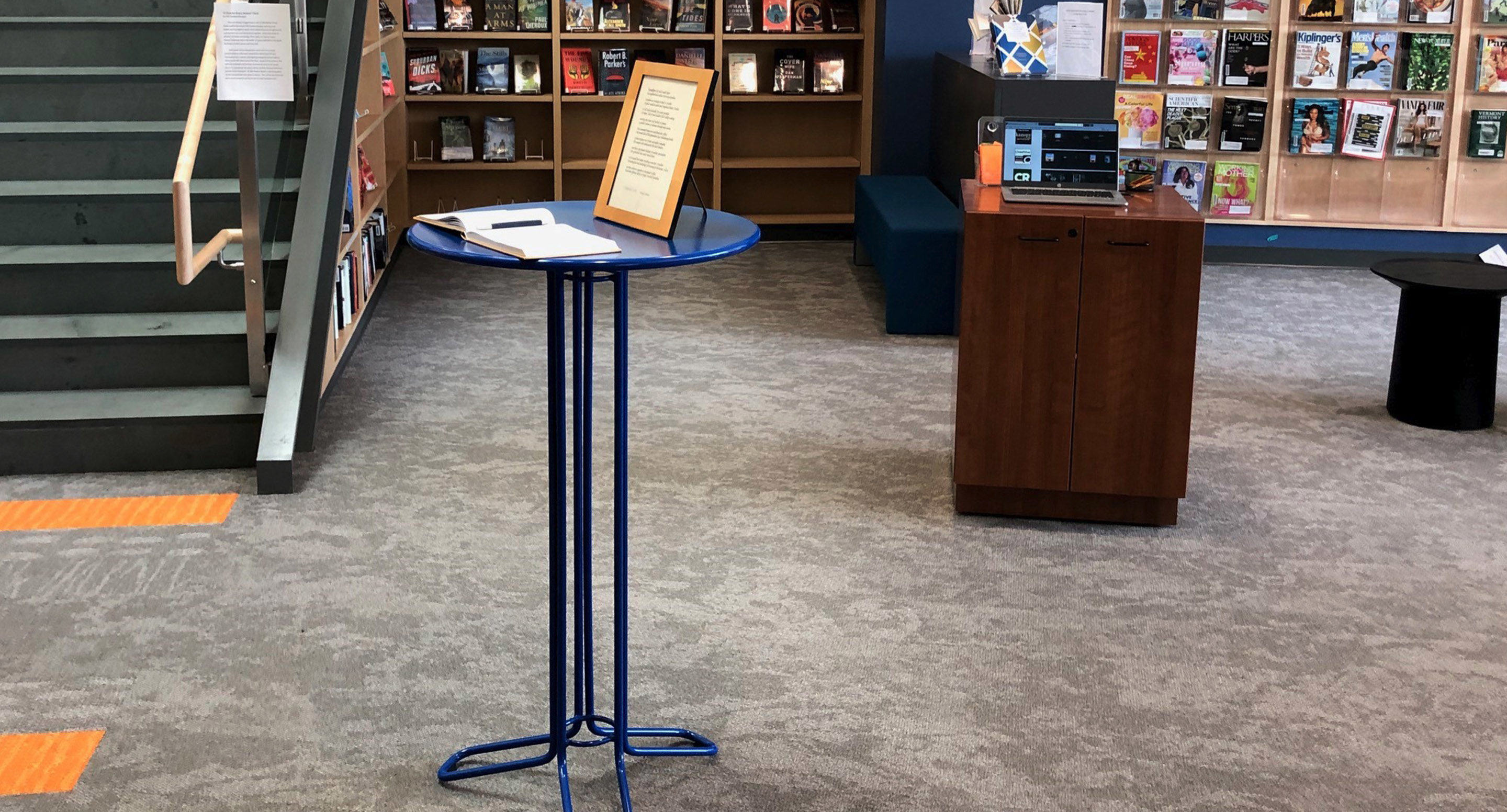 maglin_mtb-2000-kontur-bar-height-table-1 A small round table with a framed document and a notebook stands in a library near bookshelves and a staircase.