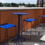 A small round blue table with two matching blue barstools sits on a tiled balcony with a wooden railing and partial view of buildings and trees in the background.