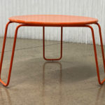 Une table ronde en métal orange avec quatre pieds courbés est posée sur un sol en béton poli devant un mur blanc texturé.