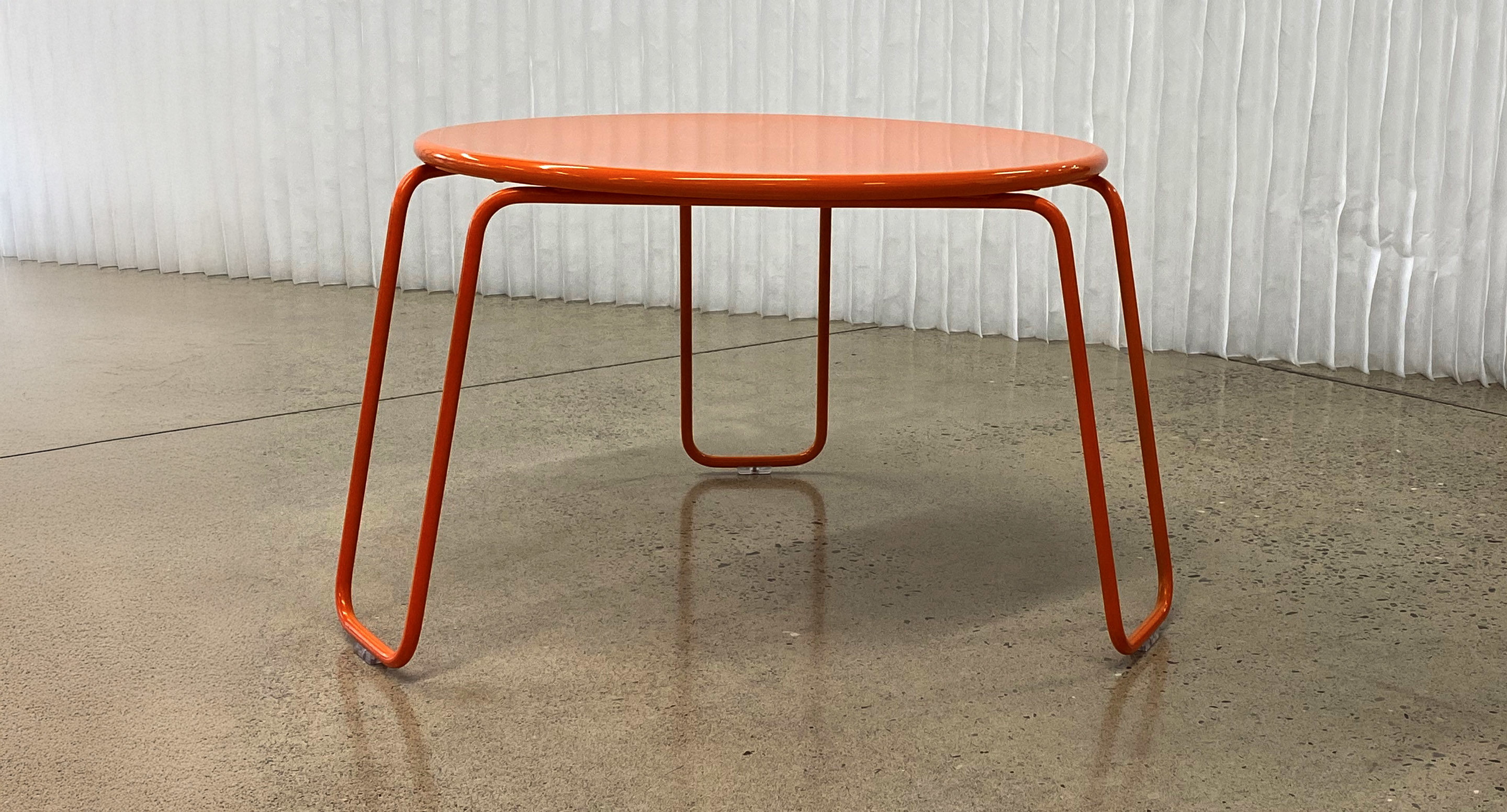 maglin_mtb-2000-kontur-lounge-table-1 Une table ronde en métal orange avec quatre pieds courbés est posée sur un sol en béton poli devant un mur blanc texturé.