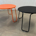 Deux tables rondes modernes aux pieds métalliques, l'une orange et l'autre noire, placées sur un sol en béton devant un mur blanc texturé.