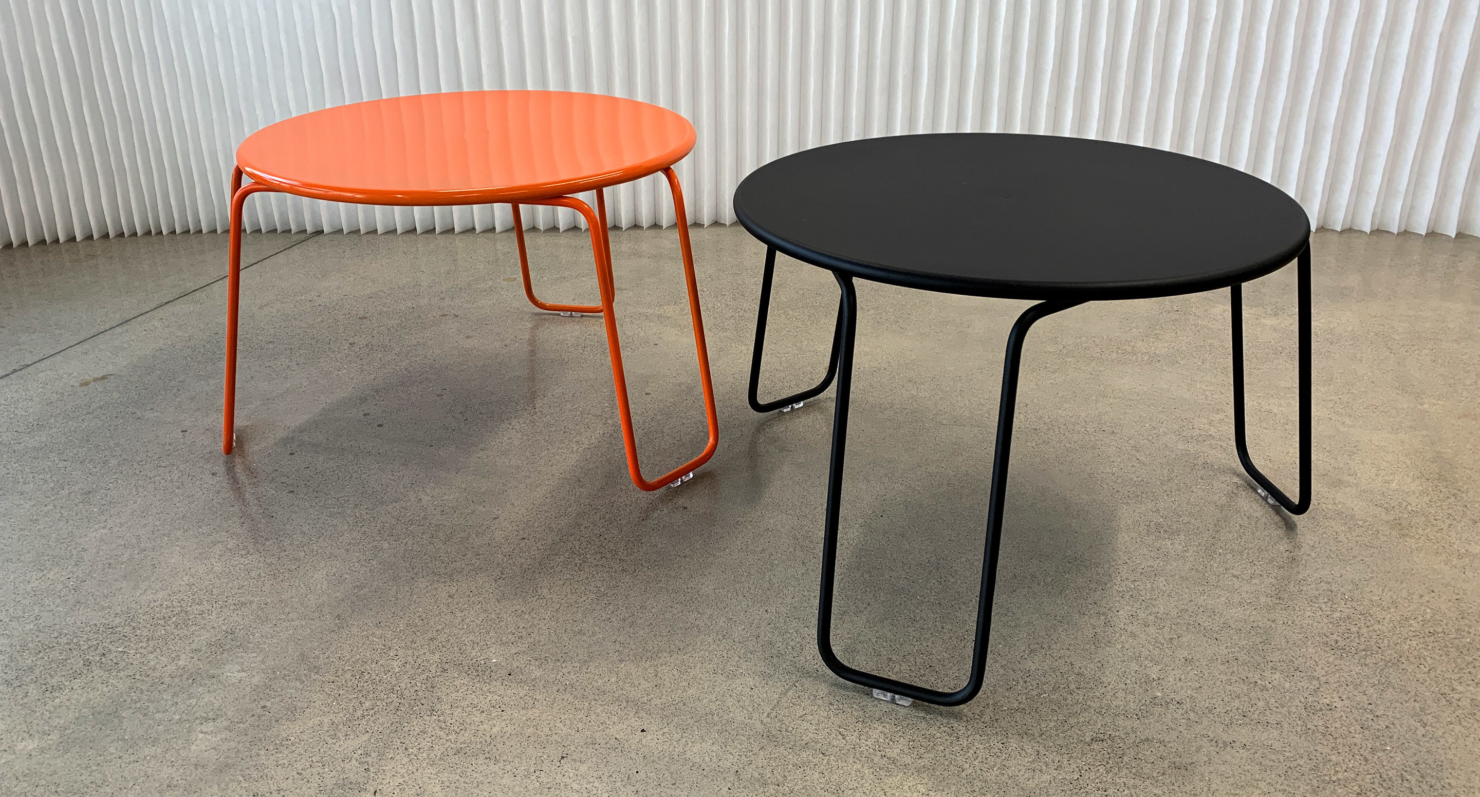 maglin_mtb-2000-kontur-lounge-table-2 Deux tables rondes modernes aux pieds métalliques, l'une orange et l'autre noire, placées sur un sol en béton devant un mur blanc texturé.