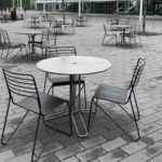 Empty metal tables and chairs are arranged in an outdoor patio area on a gray, paved surface.