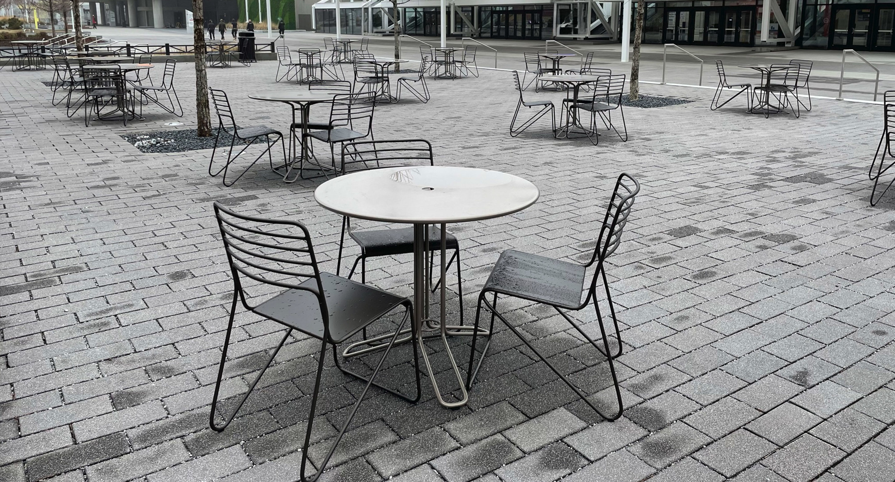 maglin_mtb-2000-kontur-tables-1 Empty metal tables and chairs are arranged in an outdoor patio area on a gray, paved surface.