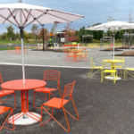 Outdoor seating area with red and yellow metal tables and chairs, large white umbrellas, and paved walkways in a park setting on a cloudy day.