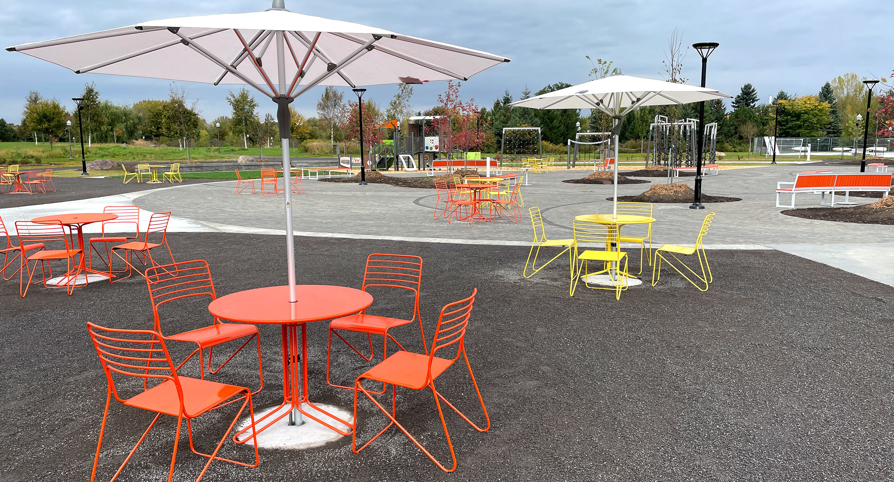 maglin_mtb-2000-kontur-tables-2 Outdoor seating area with red and yellow metal tables and chairs, large white umbrellas, and paved walkways in a park setting on a cloudy day.