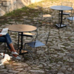 A woman sits at an outdoor café table on a stone patio, reading a book, with a white dog lying by her feet. Other empty tables and chairs are visible.