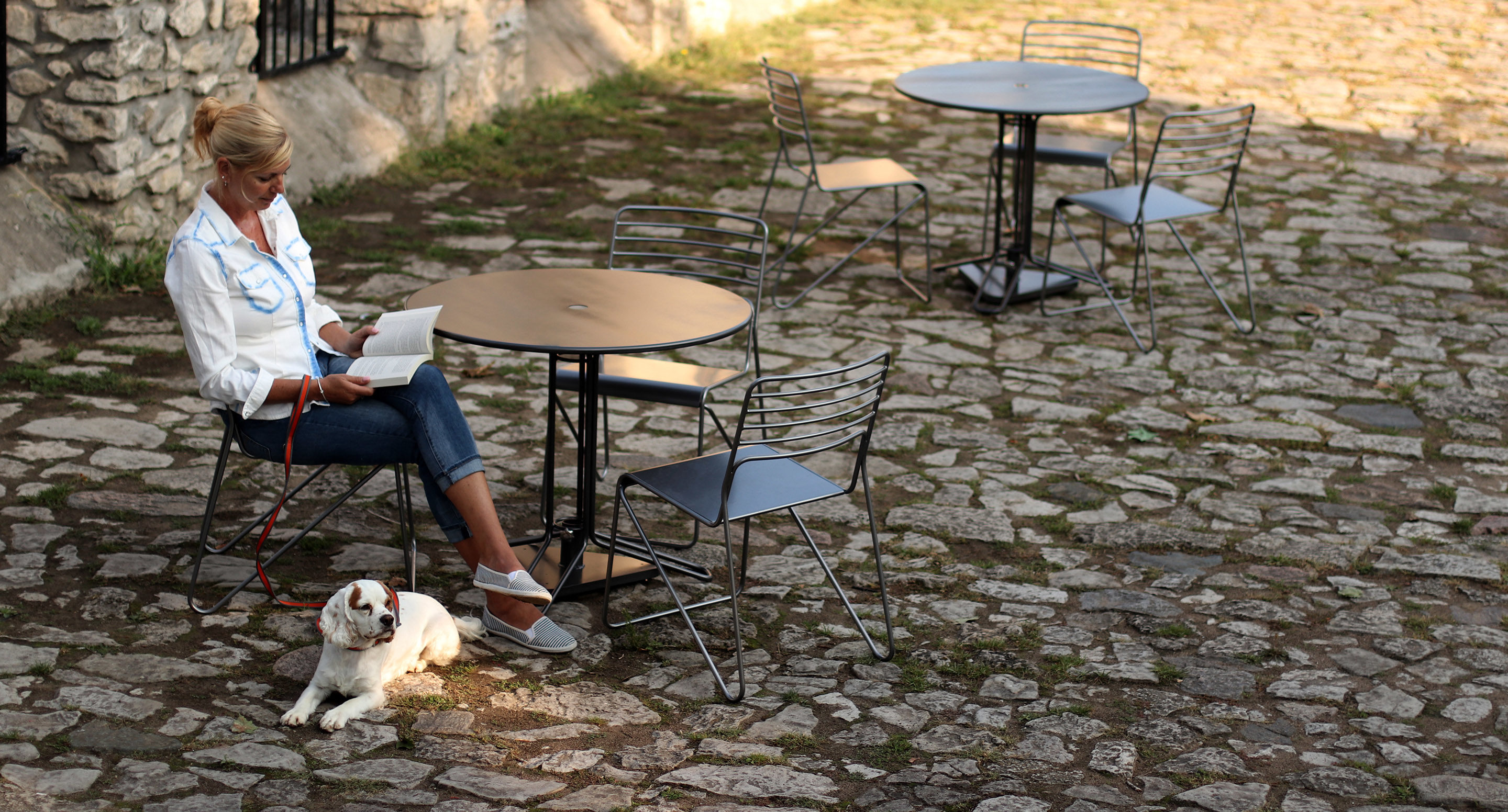 maglin_mtb-2000-kontur-tables-3 A woman sits at an outdoor café table on a stone patio, reading a book, with a white dog lying by her feet. Other empty tables and chairs are visible.