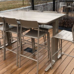 A metal table with six matching chairs is placed on a wooden deck outdoors near a grassy area and fencing.