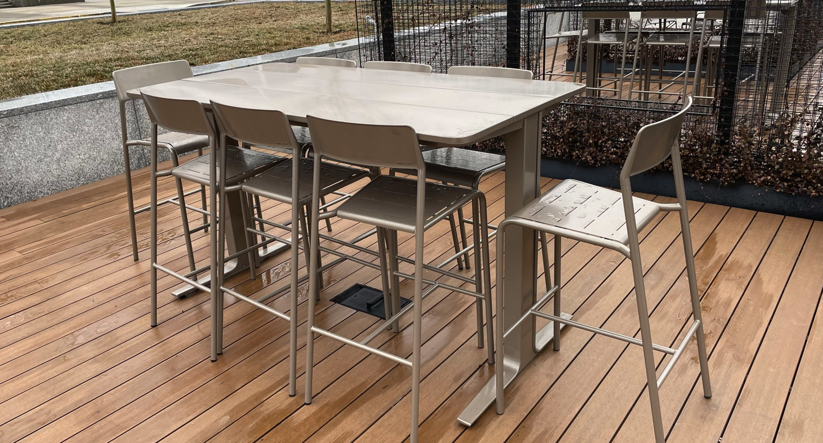 maglin_mtb-2100-ancora-tables-1 A metal table with six matching chairs is placed on a wooden deck outdoors near a grassy area and fencing.