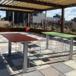 Rooftop patio with tiled floor, two tables with stools—one red, one green—under a wooden pergola, and city buildings in the background.