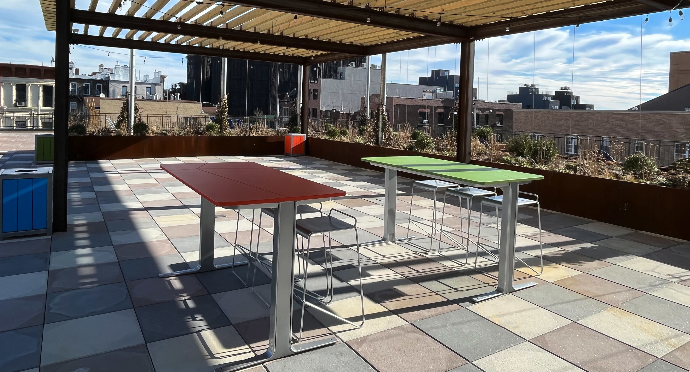 maglin_mtb-2100-ancora-tables-2 Rooftop patio with tiled floor, two tables with stools—one red, one green—under a wooden pergola, and city buildings in the background.