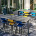Two tall tables with colorful chairs are arranged in a modern outdoor patio area next to large windows and planters with greenery.