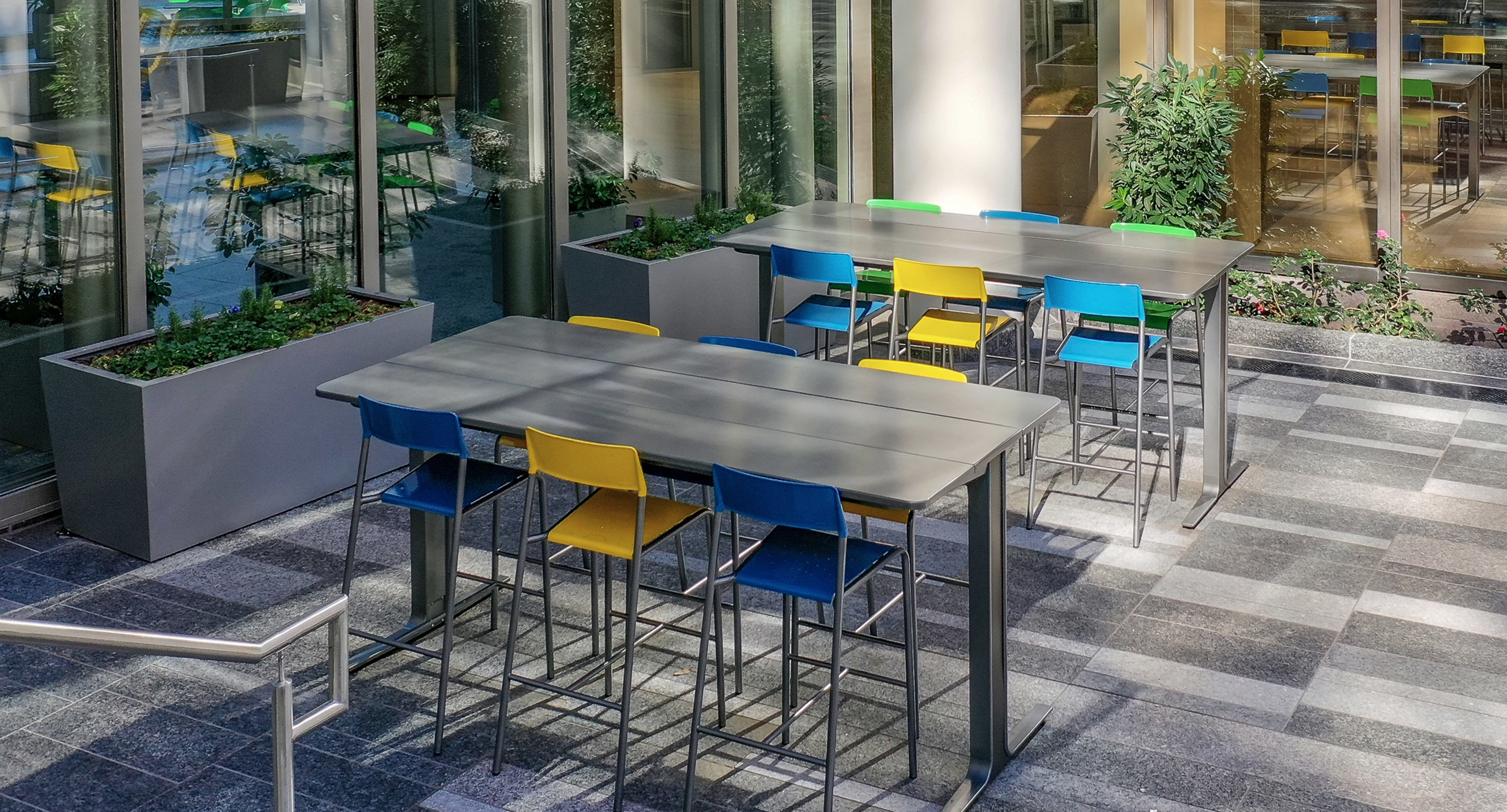 maglin_mtb-2100-ancora-tables-3 Two tall tables with colorful chairs are arranged in a modern outdoor patio area next to large windows and planters with greenery.