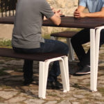 Two people sit at a wooden table with matching benches outside on a stone patio, engaged in conversation.