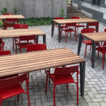 Four wooden tables with red chairs are arranged on a gray brick patio next to a modern building with striped siding and concrete walls.