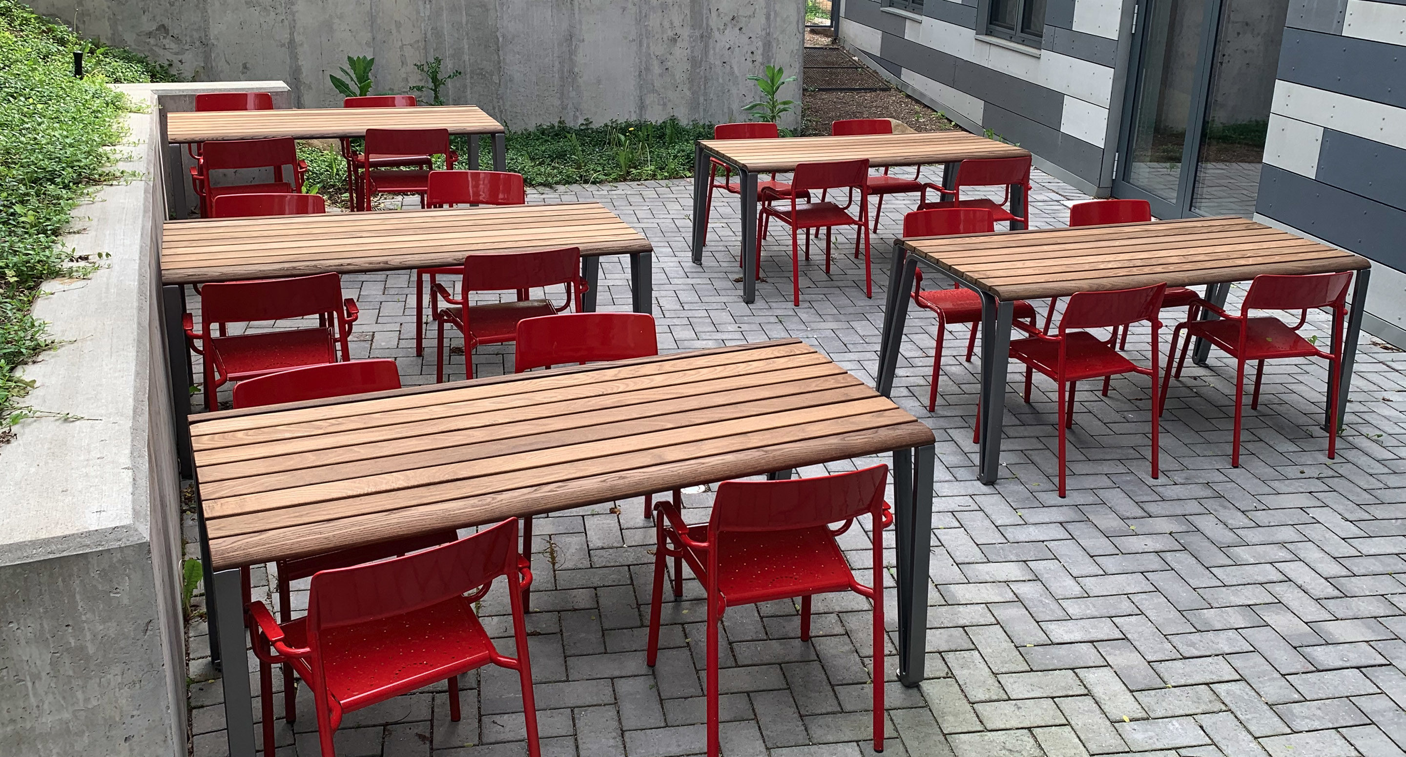 Four wooden tables with red chairs are arranged on a gray brick patio next to a modern building with striped siding and concrete walls.