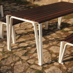 A wooden picnic table with matching benches featuring white metal legs stands on a stone-paved outdoor area in sunlight.