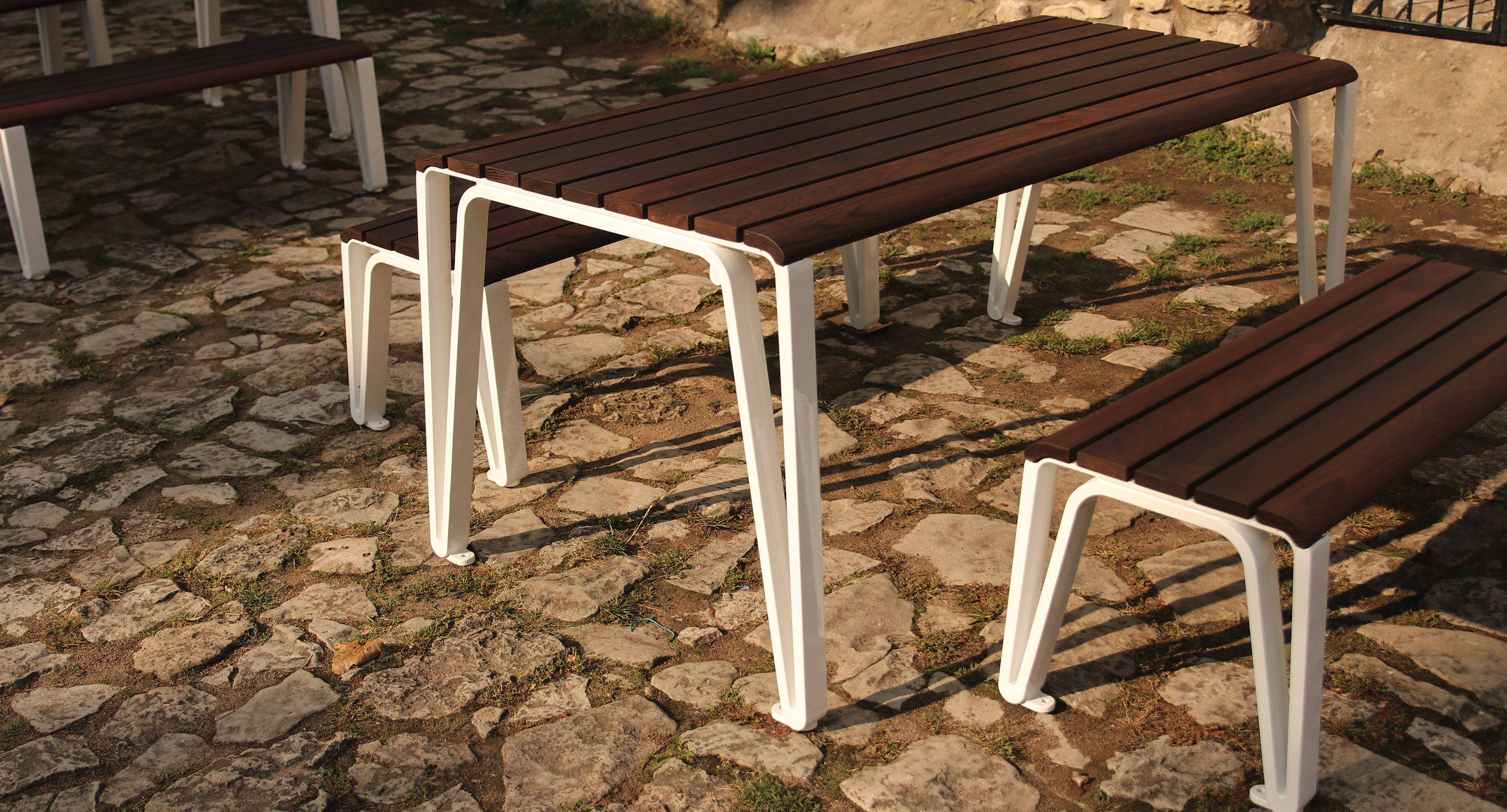 maglin_mtb-2300-iconic-tables-3 A wooden picnic table with matching benches featuring white metal legs stands on a stone-paved outdoor area in sunlight.