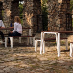 Two women sit and talk at a wooden picnic table in a stone-paved outdoor area with stone arches and additional empty tables nearby.
