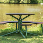 A round picnic table with built-in curved benches sits on grass near a pond, with greenery and trees in the background.