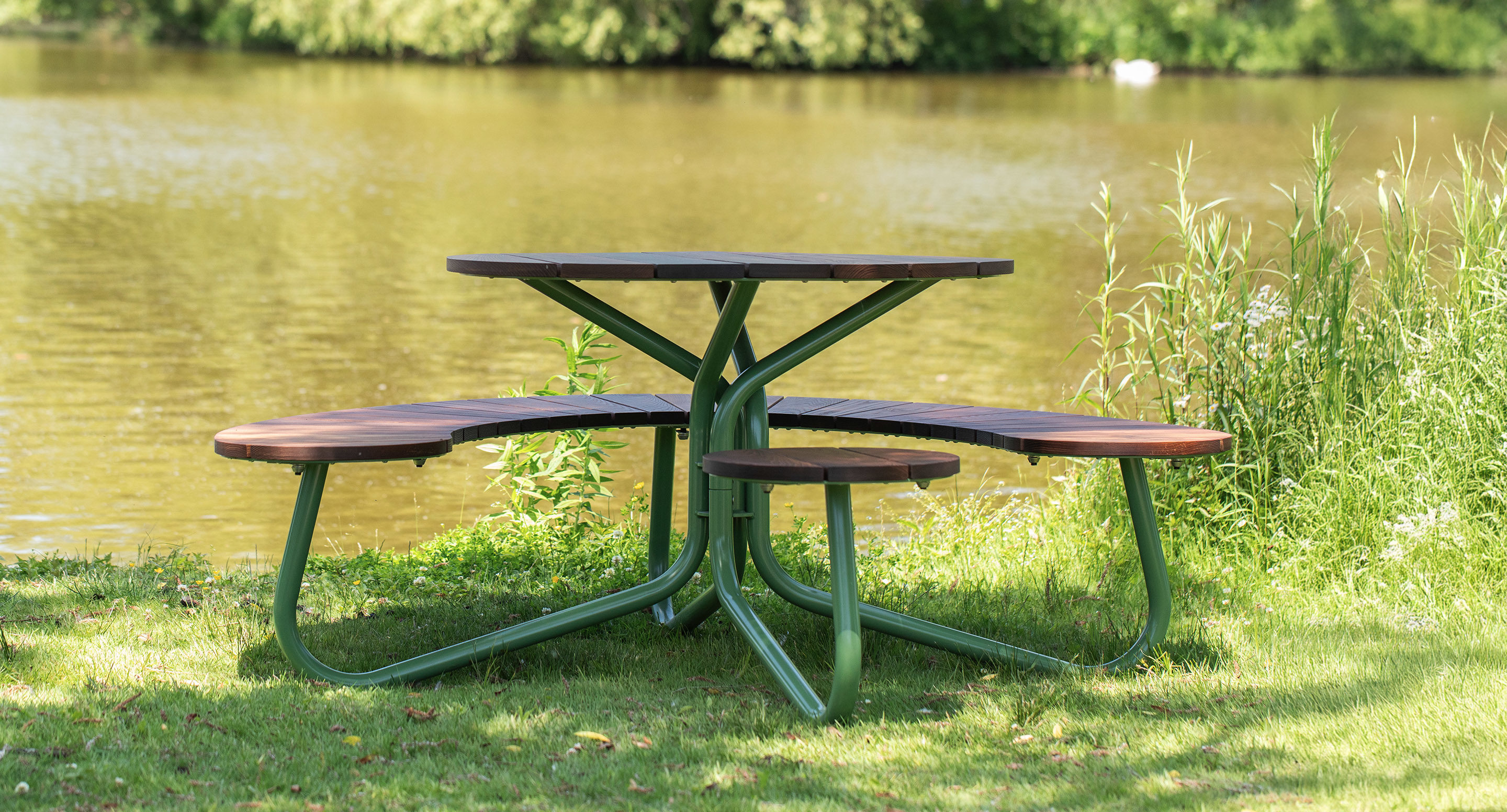 A round picnic table with built-in curved benches sits on grass near a pond, with greenery and trees in the background.