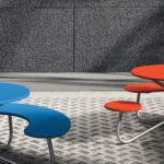 Two modern outdoor tables with curved benches, one blue and one orange, sit on a patterned stone pavement against a textured gray wall.