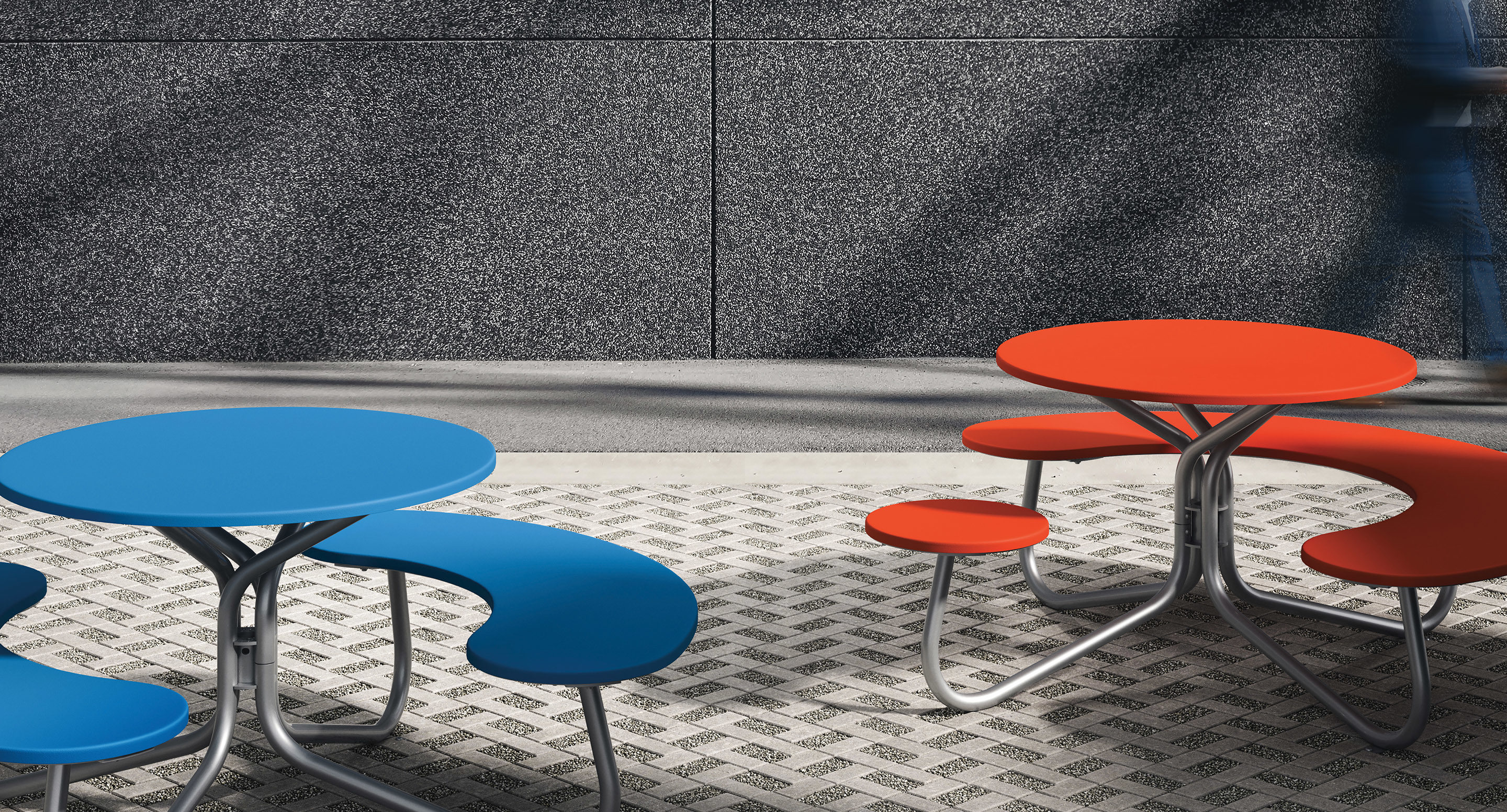maglin_mtb-2800-fava-cluster-seating-2 Two modern outdoor tables with curved benches, one blue and one orange, sit on a patterned stone pavement against a textured gray wall.