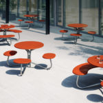 Outdoor patio with modern red round tables and curved benches, some holding glasses and a potted plant, surrounded by potted trees and garden beds.