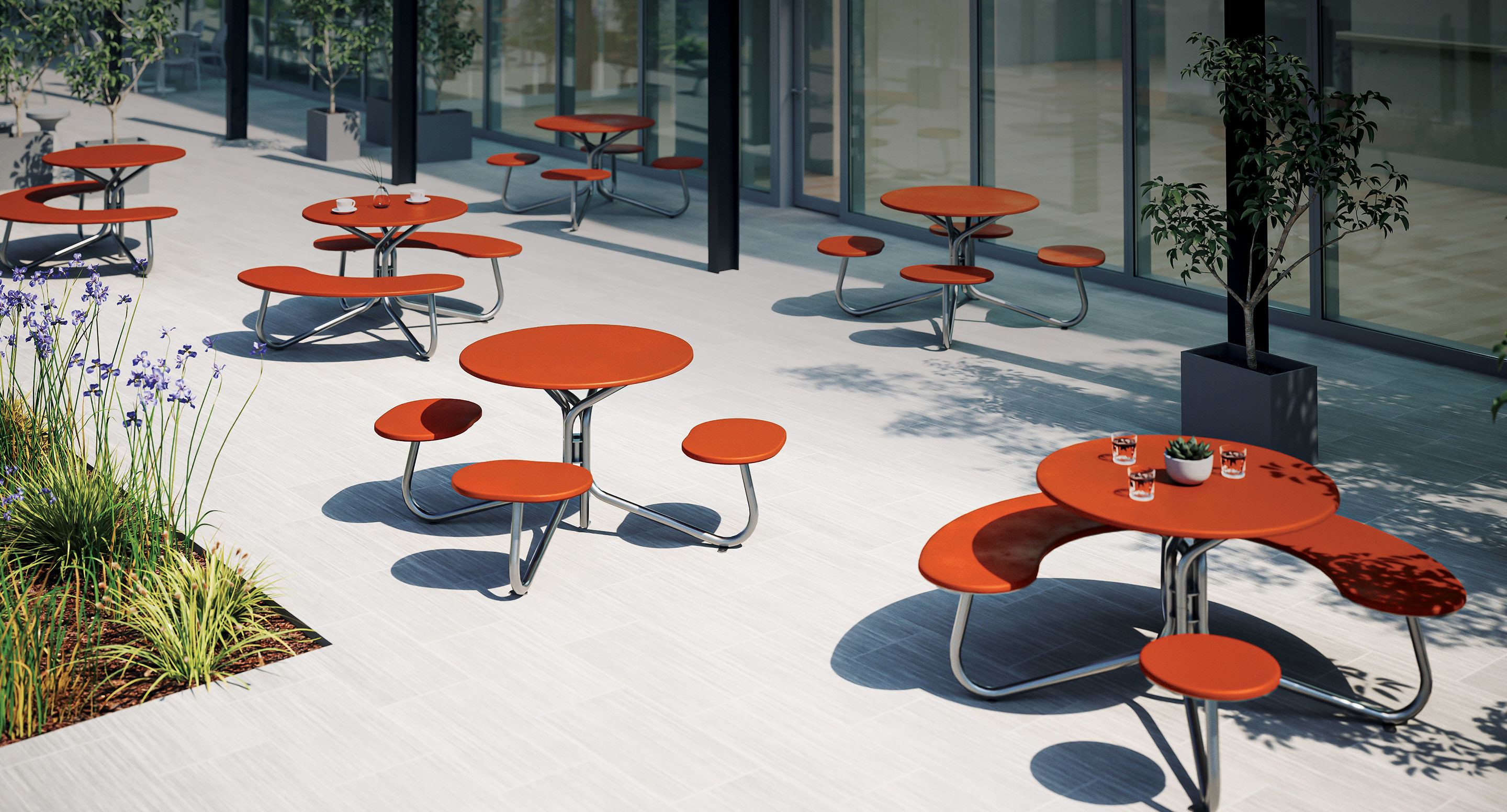 maglin_mtb-2800-fava-cluster-seating-3 Outdoor patio with modern red round tables and curved benches, some holding glasses and a potted plant, surrounded by potted trees and garden beds.