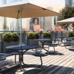 Outdoor rooftop seating area with round picnic tables, umbrellas, potted plants, and a city skyline in the background. The wooden deck is empty and sunlit.