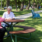 Two older adults sit and talk at a round picnic table in a park, with trees, grass, and a pond visible in the background.