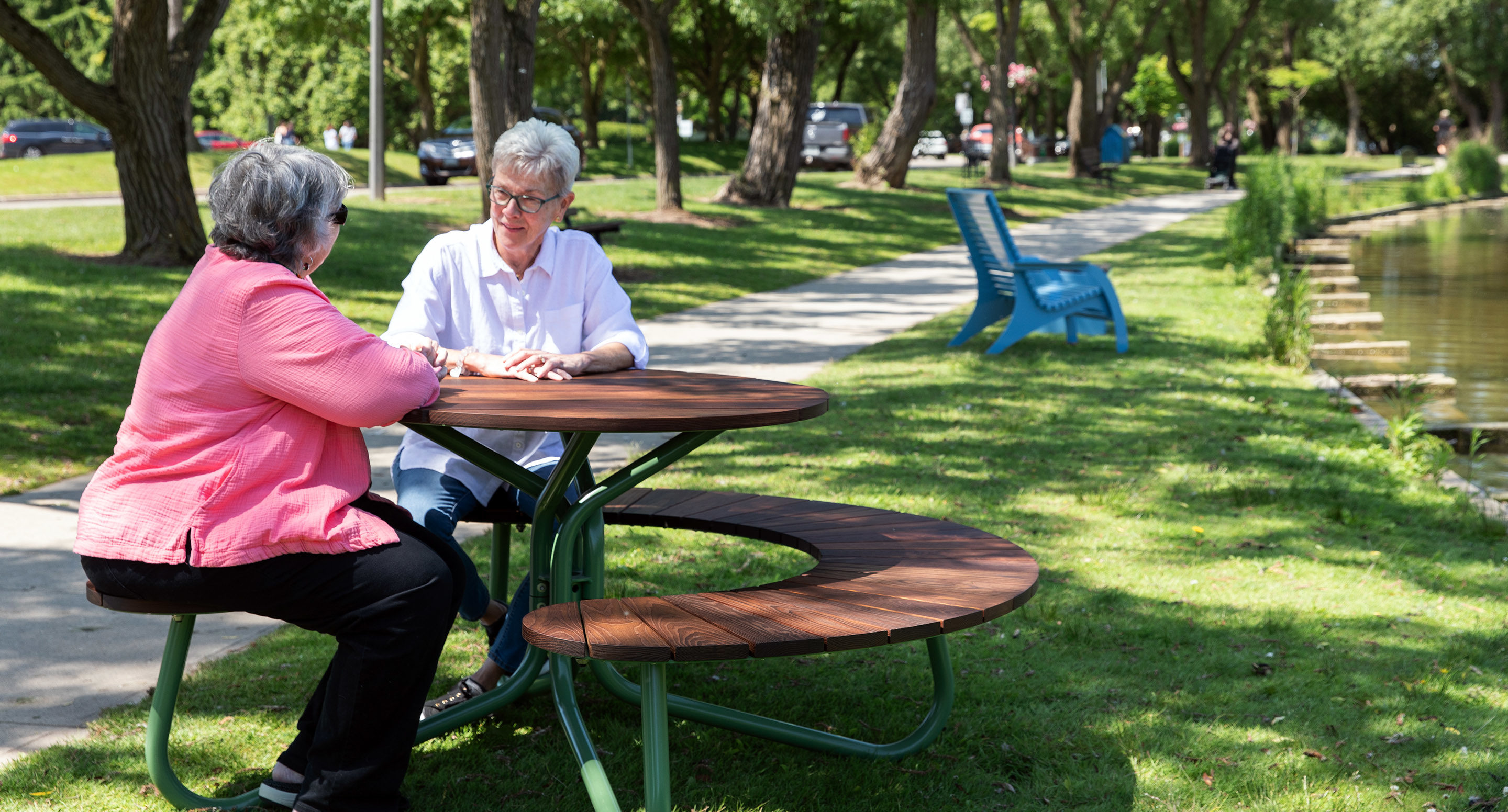 Two older adults sit and talk at a round picnic table in a park, with trees, grass, and a pond visible in the background.