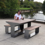 A woman sits alone at a modern outdoor picnic table near a lake, holding a drink and using her phone. Trees and water are visible in the background.