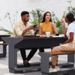 Three people sit at an outdoor bench table, talking and drinking from cups, with plants and a white brick wall in the background.