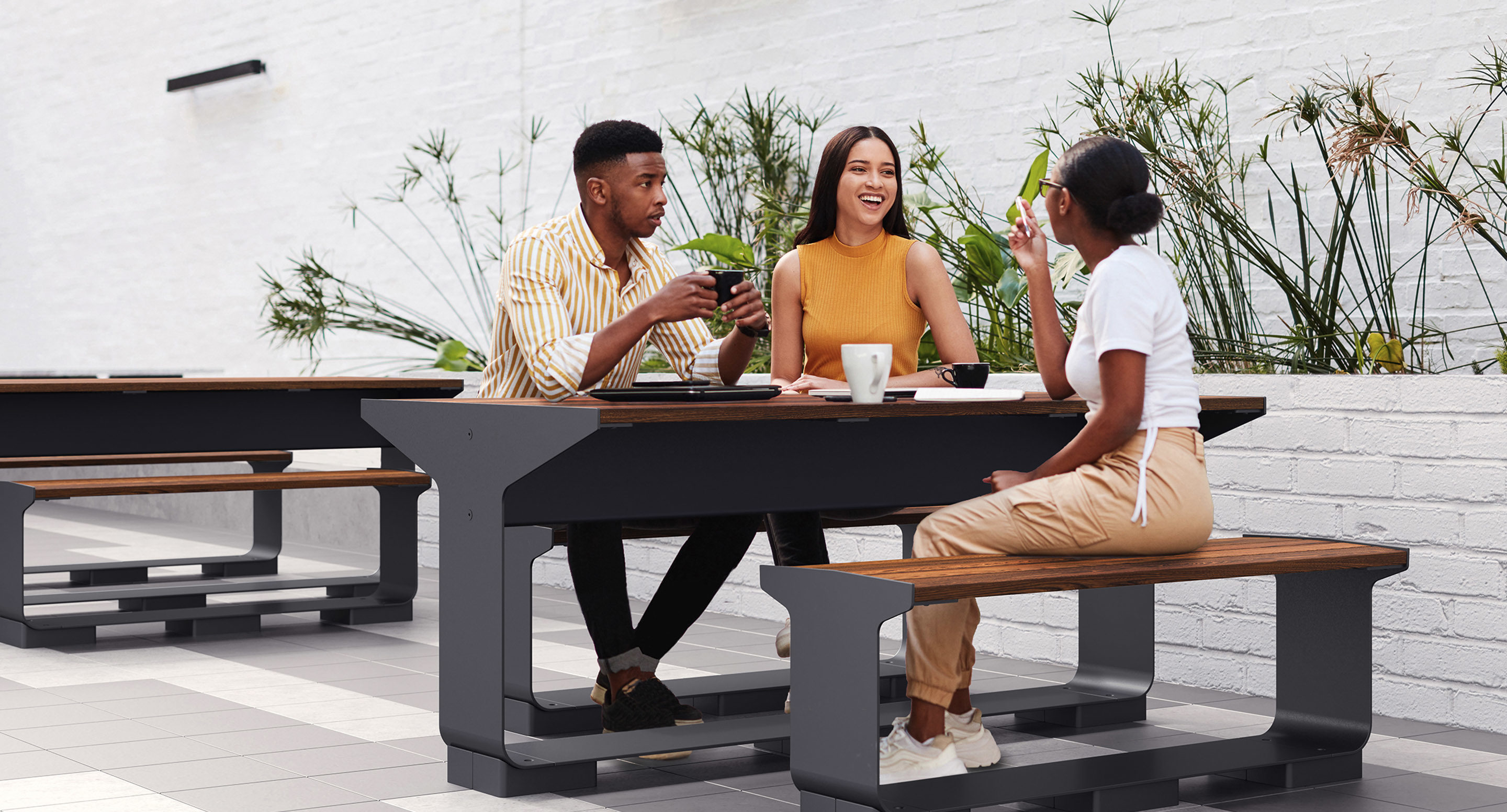 maglin_mtb-3000-l-series-tables-2 Three people sit at an outdoor bench table, talking and drinking from cups, with plants and a white brick wall in the background.