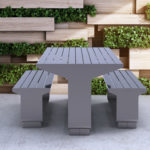 Gray metal outdoor table with benches on a concrete surface, placed in front of a decorative wall featuring wooden panels and green plants.