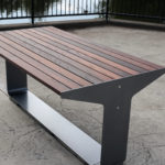 A modern wooden and metal bench is positioned on a concrete surface near a black metal railing by a body of water.