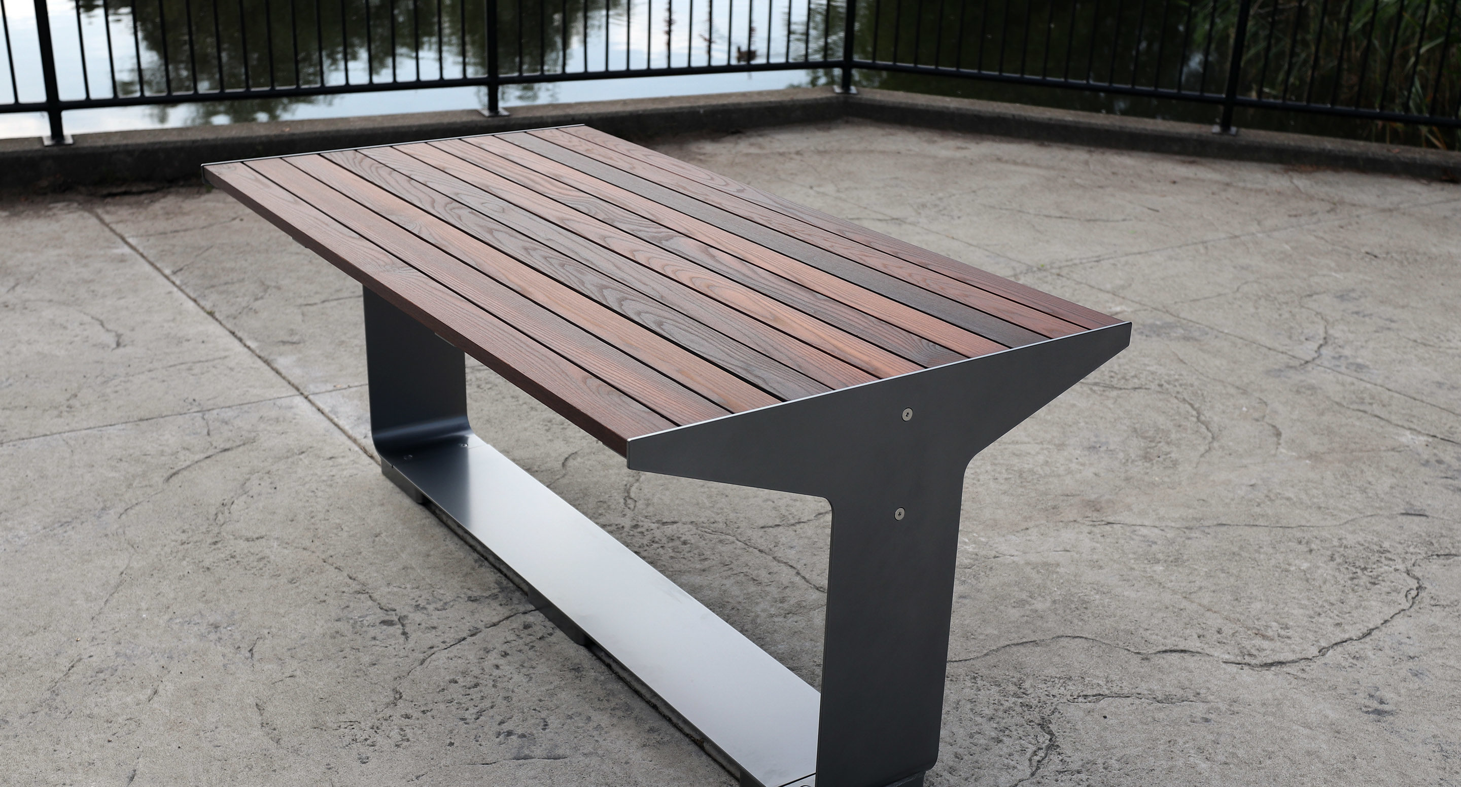maglin_mtb-3000-l-series-tables-5 A modern wooden and metal bench is positioned on a concrete surface near a black metal railing by a body of water.