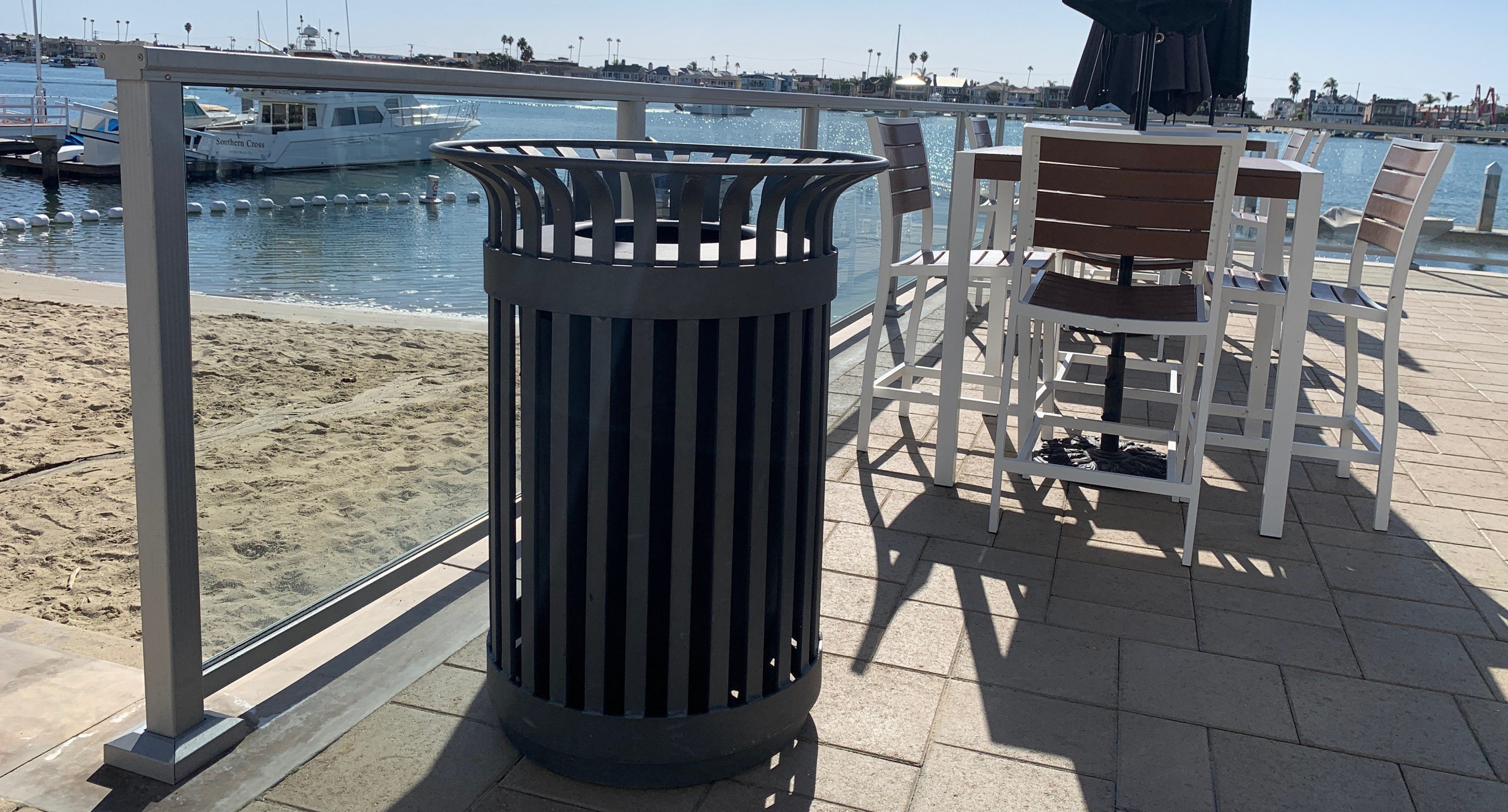 maglin_mtr-0200-trash-receptacles-0611 Une poubelle en métal noir est placée sur un patio pavé près d'une plage, avec des chaises et des tables d'extérieur blanches et un plan d'eau en arrière-plan.