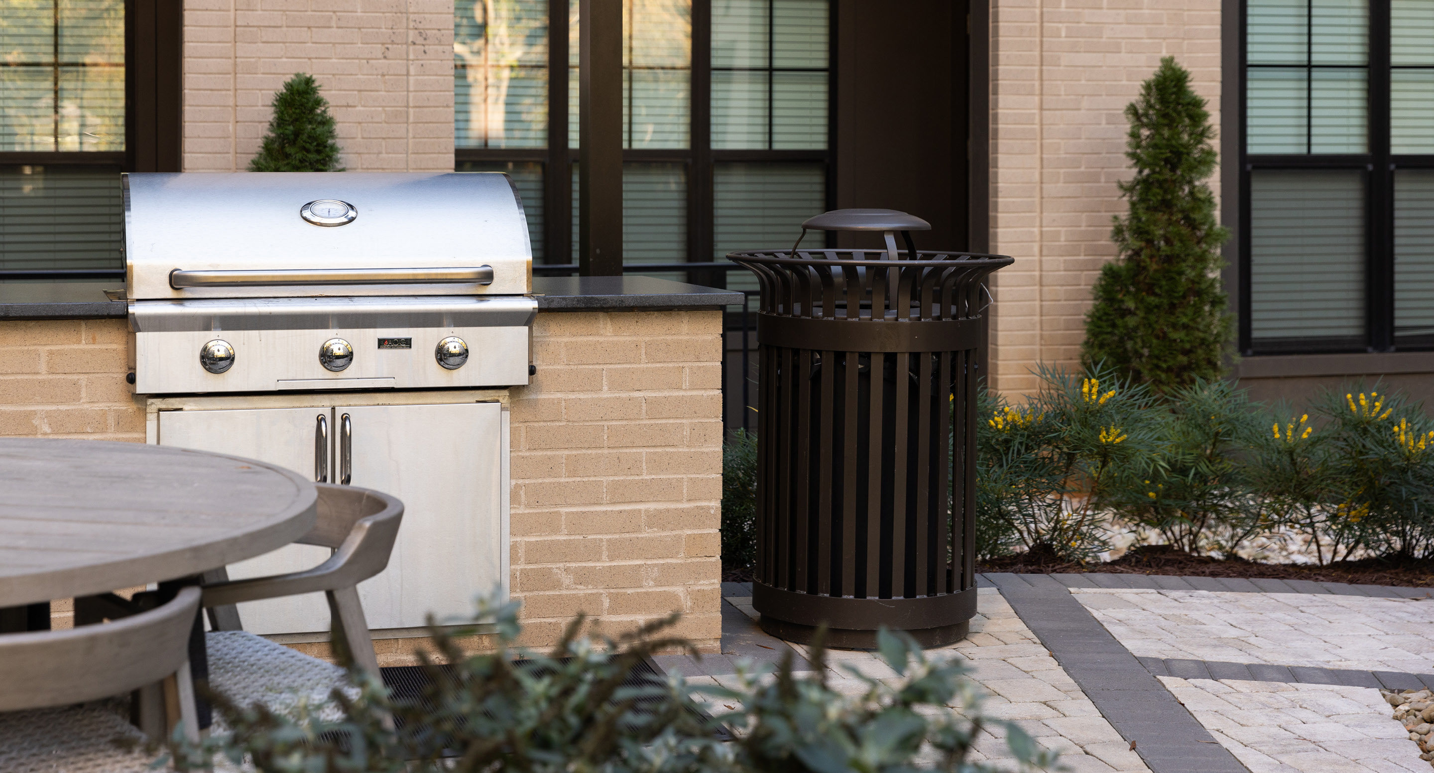 maglin_mtr-0200-trash-receptacles-1 Terrasse extérieure équipée d'un gril en acier inoxydable, d'une table ronde en bois avec des chaises, d'une poubelle en métal et d'un aménagement paysager composé de buissons et de petits arbres.